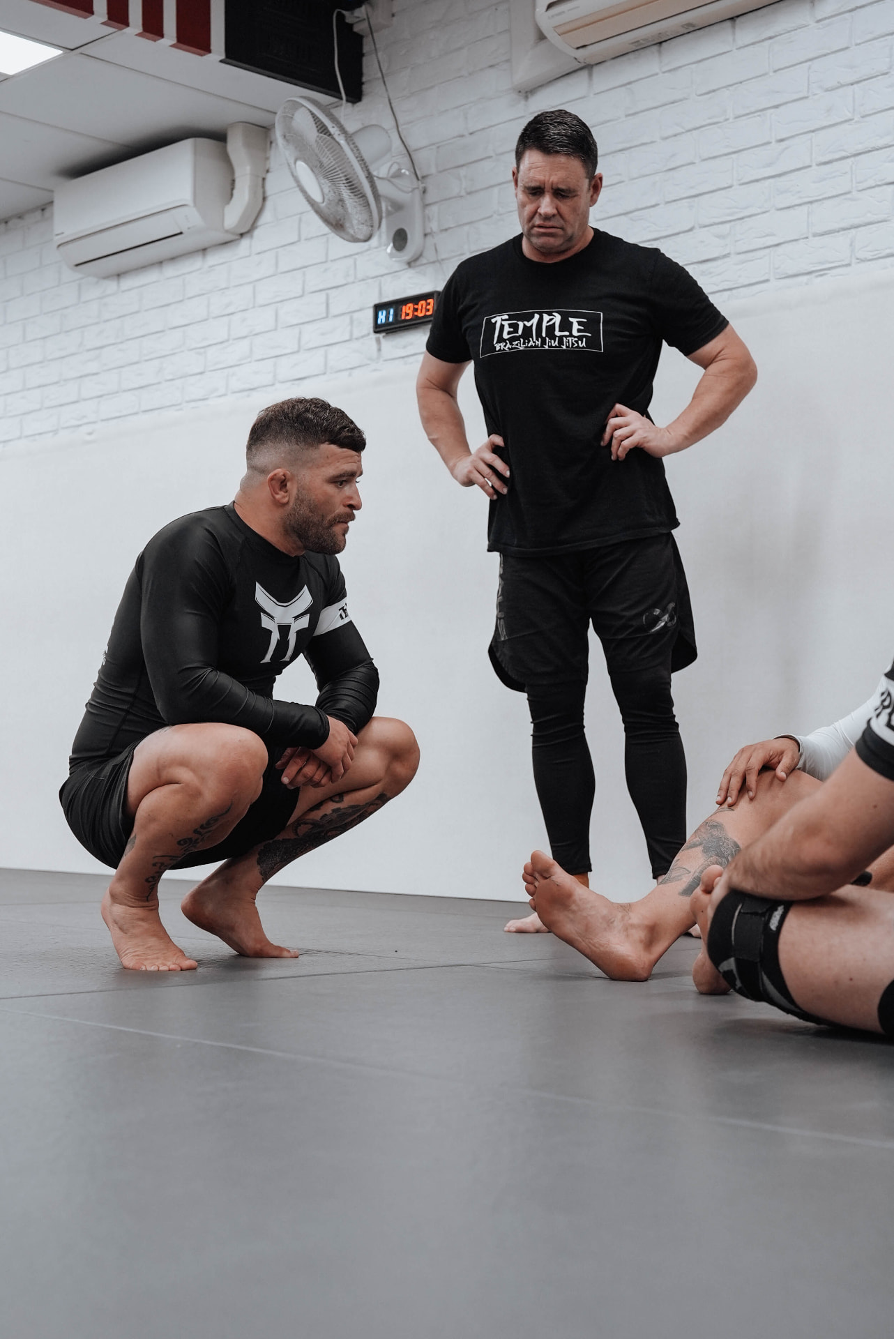 Coaches teaching an adult beginner a fundamental guard position in black gis at Temple BJJ